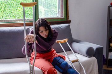 Sad young woman with crutches with a broken leg sitting on a couch.