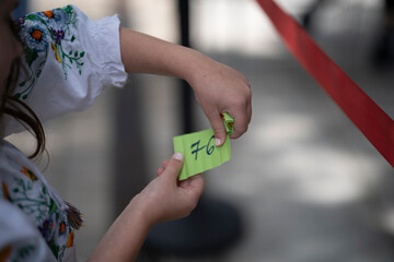 Horizontal photo of a little girl in an embroidered blouse and floral wreath holding a paper lottery ticket with the number 76, selective focus, concept of childhood curiosity and play.