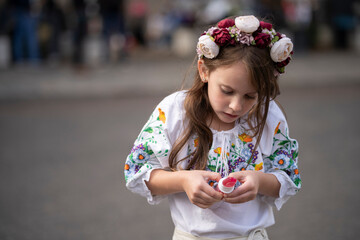 Little Ukrainian girl in an embroidered blouse and floral wreath holding a lollipop, head tilted down, focused on it with a blurred background, concept of childhood curiosity and simplicity.