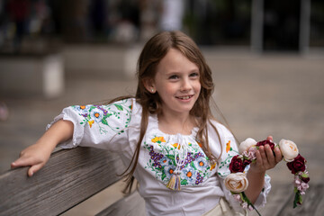 Smiling little Ukrainian girl with light brown hair in an embroidered blouse holding a floral wreath in her hands outdoors with a blurred street background, concept of childhood joy