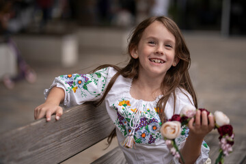 Smiling little Ukrainian girl with light brown hair in an embroidered blouse holding a floral wreath in her hands outdoors with a blurred street background