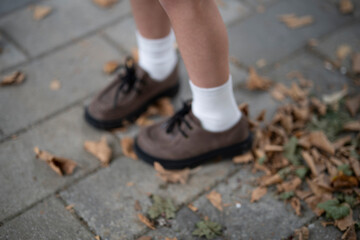 Blurred photo of children’s stitched leather shoes with laces and white socks on cobblestone ground with fallen leaves, copy space, concept of childhood and autumn.