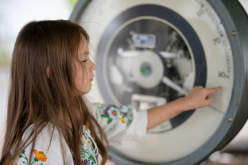 Side view of a long-haired girl pointing at the dial of large vintage scales, face visible, concept of curiosity and childhood learning.