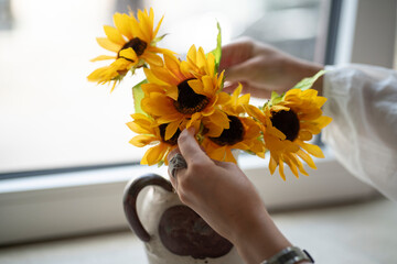 Female hands arranging a composition of sunflowers in a vase on a windowsill, concept of home comfort and creativity.