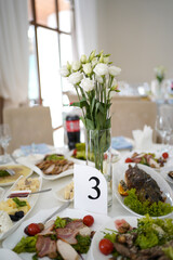 Festive table with various dishes, salads, meat and fish, table setting with flowers in a vase and table number.