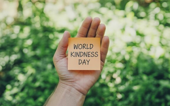 A hand holding an orange note with world kindness day written on it in front of a green background