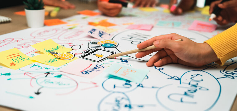 A close-up view of hands involved in brainstorming with sticky notes on chart paper, showcasing teamwork, creativity, and idea generation in a collaborative workspace. SACTR - Powered by Adobe