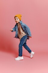 Young boy wearing an orange beanie and denim jacket is playfully walking against a pink background,...