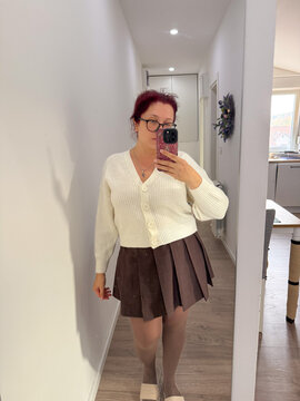 Woman in white cardigan and brown skirt taking mirror photo. Light tones, home interior, and balance of color and texture.