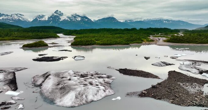 Footage above the river with melting ice boulders. Campers are at the waterfront on the other side. Mountain range covered with clouds at tops at backdrop. Alaska, USA.