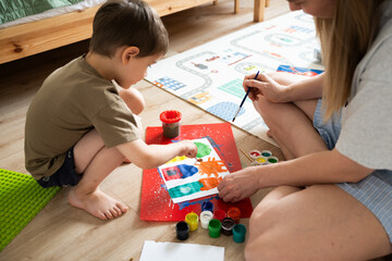 Child is painting alongside mother in a vibrant indoor space, surrounded by colorful art supplies and playful atmosphere