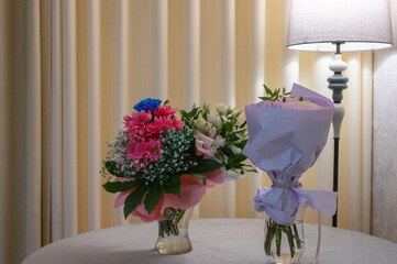 Two floral bouquets in glass vases on a table with soft lighting and a lamp in the background.