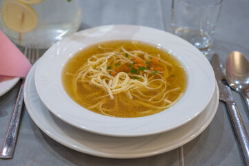 Clear broth soup with noodles, diced vegetables, and herbs served in a white bowl on a table setting.