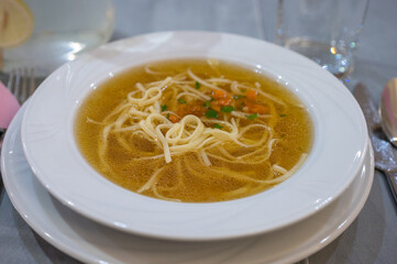 Clear broth soup with noodles, diced vegetables, and herbs served in a white bowl on a table setting.