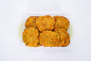 A close-up image of homemade vegetable fritters, neatly arranged in a transparent plastic container. The golden-brown patties appear crispy on the outside with visible bits of herbs and vegetables.
