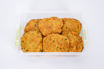 A close-up image of homemade vegetable fritters, neatly arranged in a transparent plastic container. The golden-brown patties appear crispy on the outside with visible bits of herbs and vegetables.
