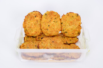 A close-up image of homemade vegetable fritters, neatly arranged in a transparent plastic container. The golden-brown patties appear crispy on the outside with visible bits of herbs and vegetables.
