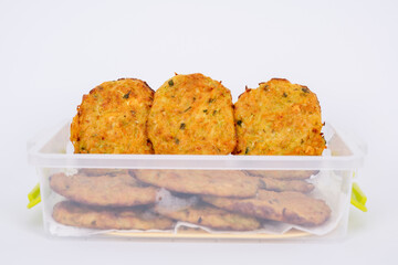 A close-up image of homemade vegetable fritters, neatly arranged in a transparent plastic container. The golden-brown patties appear crispy on the outside with visible bits of herbs and vegetables.
