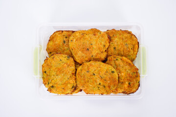 A close-up image of homemade vegetable fritters, neatly arranged in a transparent plastic container. The golden-brown patties appear crispy on the outside with visible bits of herbs and vegetables.
