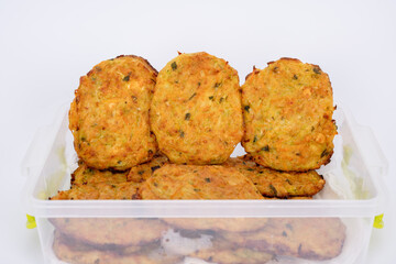 A close-up image of homemade vegetable fritters, neatly arranged in a transparent plastic container. The golden-brown patties appear crispy on the outside with visible bits of herbs and vegetables.
