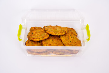 A close-up image of homemade vegetable fritters, neatly arranged in a transparent plastic container. The golden-brown patties appear crispy on the outside with visible bits of herbs and vegetables.
