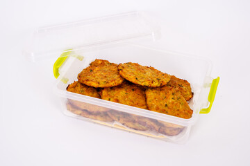 A close-up image of homemade vegetable fritters, neatly arranged in a transparent plastic container. The golden-brown patties appear crispy on the outside with visible bits of herbs and vegetables.
