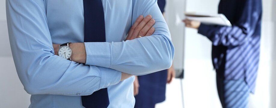 Business professional in a light blue shirt and dark tie standing with arms crossed, projecting confidence and corporate acumen in an office environment