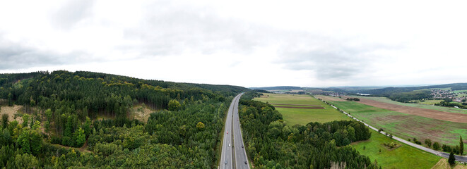 Aerial panoramic view of a straight road stretching between dense forest and open fields. The scene symbolizes travel, connection, and the balance between human routes and wild nature.