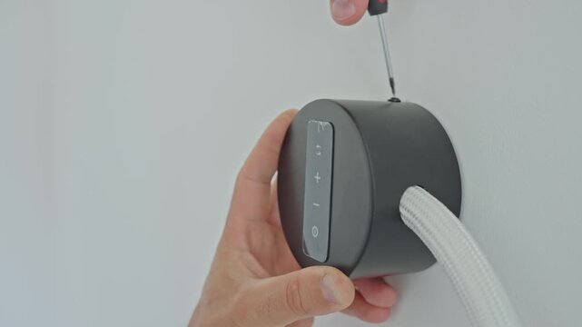 Electrician using screwdriver to install black wall-mounted device with a flexible white cable. Concept of electrical installation and home improvement