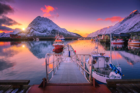 Fishing boats in harbor at sunset with snowy mountains and colorful sky in Lofoten Islands, Norway. Scenic waterfront with pier, vessels, reflection in calm water, and Arctic winter landscape