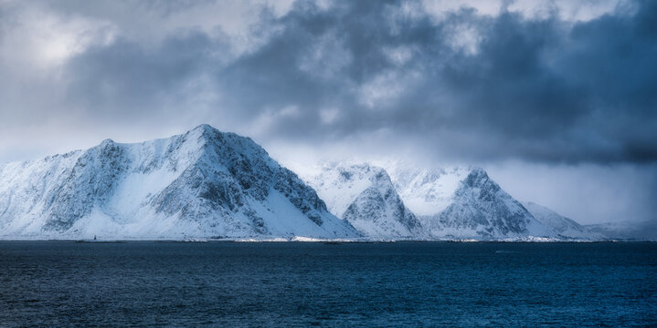 Majestic snow-capped mountains rise from the deep blue ocean under dramatic cloudy sky. Winter landscape with snowy rocks, overcast sky and sea in the Lofoten islands, Norway. Nature. Arctic scenery - Powered by Adobe