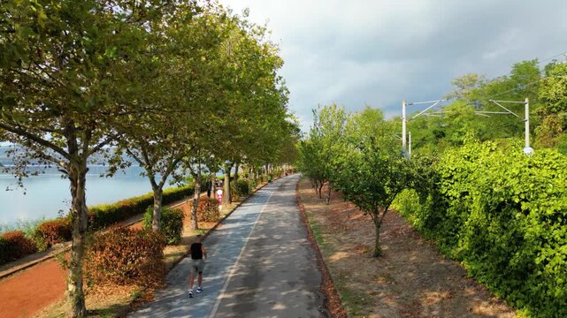 Active senior man (50-60s) jogging on a lakeside promenade for fitness, with a high-speed train and motorway visible in the background. Ascending aerial drone shot (crane up).