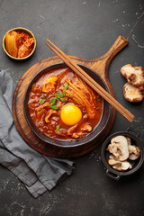 Top view of spicy kimchi jjigae soup in a black bowl with egg yolk, enoki, shiitake, tofu and green onions, with chopsticks and cooking ingredients, cuisine of Korea