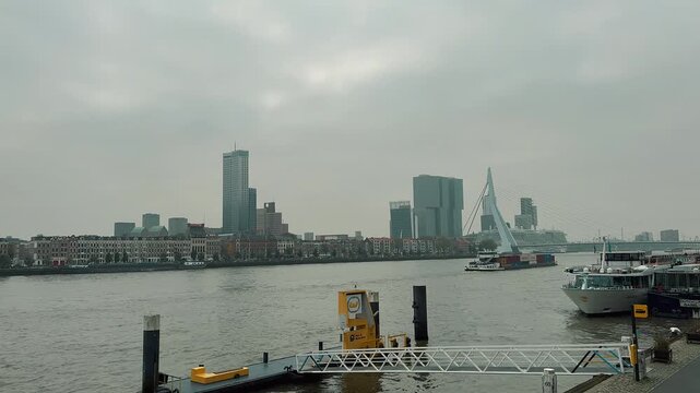 A long cargo container barge travels along the Nieuwe Maas river in Rotterdam, Netherlands, against a cloudy sky. The vessel carries international shipping containers past the impressive city skyline 