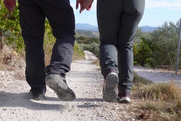 Close-up of two hikers legs and boots walking together on a rocky trail between vineyards and olive groves under clear blue sky.