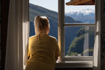 A blonde woman with a ponytail looks out a window at a stunning Norwegian fjord and snow-capped mountains.