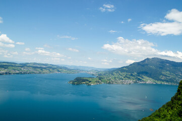 Naklejka premium A winding hiking trail meanders through a vibrant green alpine meadow, inviting adventurers to explore the stunning Swiss landscape. This beautiful scene is framed by clusters of lush trees and tradit