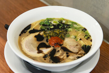 Bowl of rich pork tonkotsu ramen with black garlic oil, spicy paste, pickled greens and green onions in savory broth on wooden table