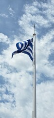 Greek flag flutters against blue sky with white clouds.