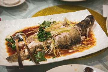 Traditional Chinese steamed fish garnished with ginger, scallions, and cilantro in soy-based sauce served on large white platter with banquet spoons