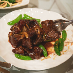 Cantonese style stir-fried beef cubes with sugar snap peas and onions in glossy brown sauce, plated on white dish with fork picking up a piece