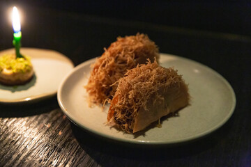 Crispy kataifi rolls made with shredded phyllo dough, served on round plate under soft ambient lighting with candle-lit pistachio topped sweet in background