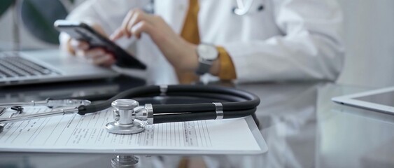Close-up of doctor's desk with stethoscope and clipboard. Physician is using a laptop computer at the background. Medicine concept