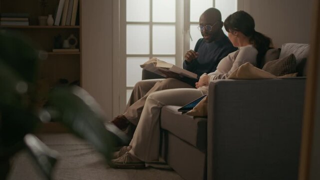 African American man reading book out lout to his pregnant wife, listening to him attentively and stroking her stomach while sitting next to him on couch in cozy living room