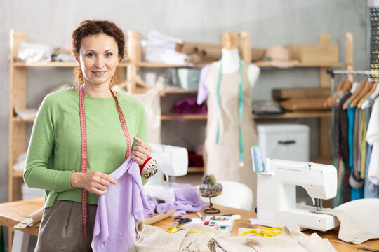 Smiling female tailor with measuring tape around neck and pincushion with colorful pins on hand standing in sewing studio, posing during break at work surrounded by tools, fabrics and garments on rack