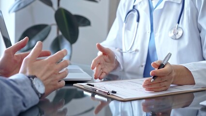 Doctor woman wearing white medical coat is consulting patient in clinic office while both are sitting at the glass desk. Physician is listening and making notes. Medicine concept