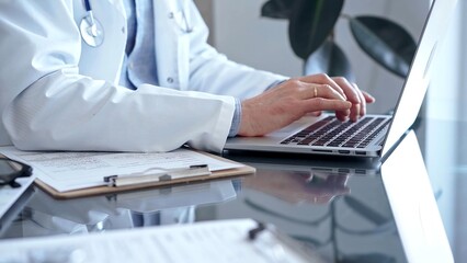 Doctor man with blue shirt and medical coat is using laptop computer in clinic office while sitting at the glass desk. Medicine concept