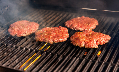 Close up of four raw hamburgers cooking on a grill with smoke and flame
