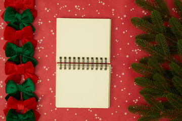 Open blank notebook in the center with Christmas tree branches and festive ribbons on a red background, holiday flat lay composition.