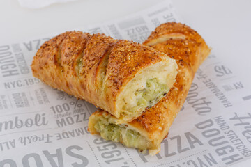 Freshly baked savory bread roll with green herb and cheese filling, topped with sesame seeds. Served on branded pizza paper with print. Crispy crust and soft interior visible in cross-section.